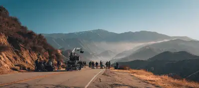 A film crew sets up equipment on a mountain road while filming in Vietnam, with people gathered around cameras and lighting gear. Rolling hills and distant mountains are visible under a clear blue sky.