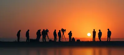 Silhouettes of people and film equipment are visible against a vibrant orange sunset sky, capturing the essence of filming in South Africa, with the sun near the horizon and a calm reflective surface in the foreground.