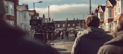 A person in a jacket stands on a residential street filming in Norway, near a film camera setup, with houses lining both sides and a distant group of people visible under a cloudy sky.