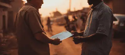 Two people stand facing each other outdoors at sunset, holding and looking at a document or clipboard together while filming in Switzerland, with blurred buildings and people in the background.