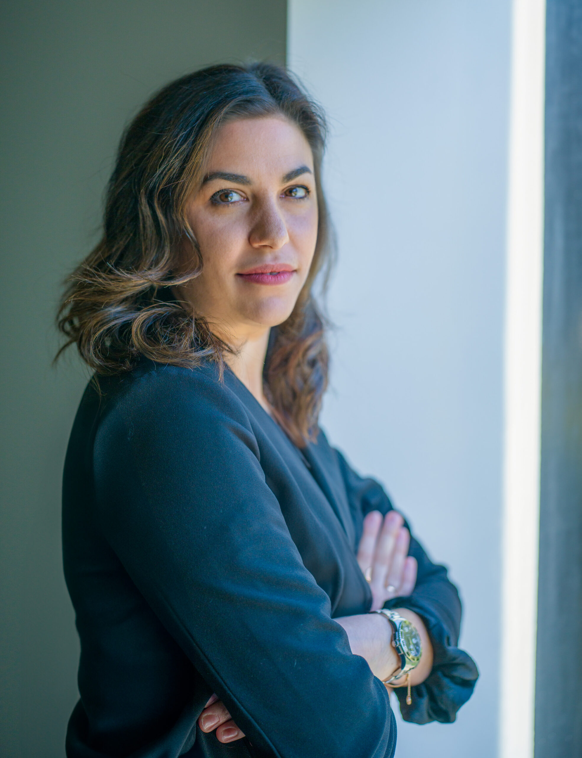 A woman with wavy brown hair, wearing a dark blouse and a watch, stands with her arms crossed and looks at the camera while leaning against a wall near a bright window.