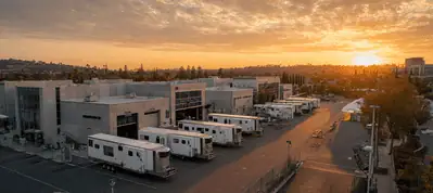 A row of white trailers is parked outside large studio buildings at sunset, with a warm orange sky and scattered clouds in the background, capturing the atmosphere of filming in Moscow.