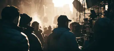 A film crew and actors stand on a city street at dawn or dusk in Austin, their silhouettes outlined against a bright, hazy sky. Filming equipment is in use as urban buildings and scaffolding frame this scene of filming in Austin.