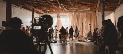 A film crew works on a set in a large studio with exposed beams, lighting equipment, and cameras, preparing for filming in Wellington near a window with beige curtains.