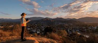A person wearing safety gear stands on a rocky hill at sunset, holding a clipboard and surveying a town below—perfect for filming in Austin—with mountains and a partly cloudy sky in the background.