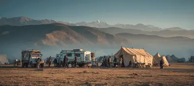 A campsite with several tents and vehicles sits on dry ground, with people gathered outside. Mountains and misty hills rise in the background under a clear sky, capturing the essence of filming in the Canary Islands.