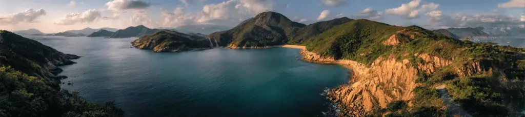 A panoramic view of a coastal landscape—perfect for filming in Hong Kong—features calm blue water, rocky cliffs, and lush green hills under a partly cloudy sky.