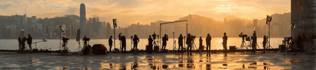 A film crew with cameras and equipment is silhouetted against a city skyline at sunset, filming in Hong Kong on a waterfront set with mountains and tall buildings in the background.