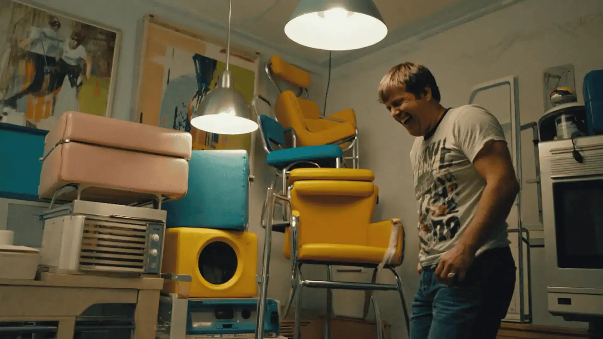 A man stands smiling in a room filled with colorful retro appliances and stacked yellow and blue chairs, illuminated by hanging lights—reminiscent of how the best comedy is filmed, with vibrant sets full of character.