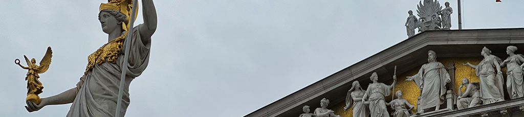 A large statue of a robed woman holding a staff and a small winged figure stands beside a building in Austria, adorned with golden and white classical statues and intricate relief carvings.