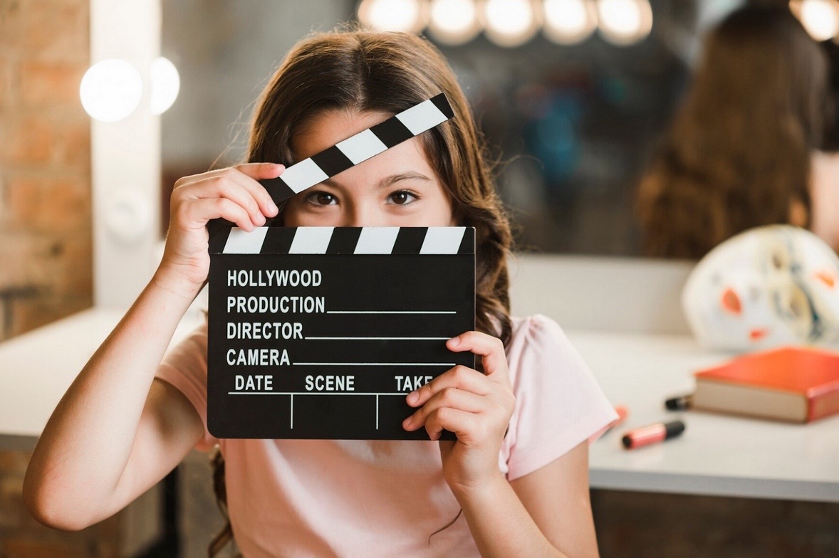 A young girl holds a movie clapperboard in front of her face, showing just her eyes—a playful nod to the art of film review. She stands in a dressing room with a mirror, lights, makeup, and a book in the background.