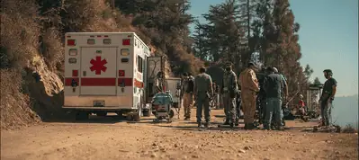A group of people, including emergency responders, stand near an ambulance on a dirt road surrounded by trees and hills in the Canary Islands, with medical equipment set up nearby under clear skies during filming.