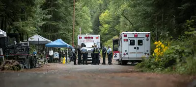 Several emergency responders stand near two ambulances on a wooded road, with tents and equipment set up nearby—possibly for filming in Norway—surrounded by tall green trees and dense foliage.