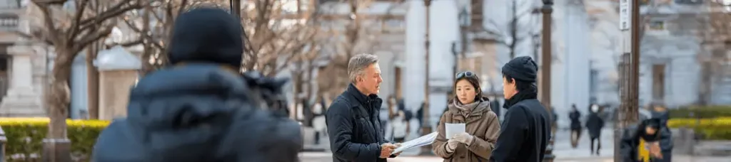 A group of three people dressed in winter clothing stands outside in a city square, having a conversation and holding papers—possibly preparing for filming in Japan. Leafless trees and blurred buildings are visible in the background.