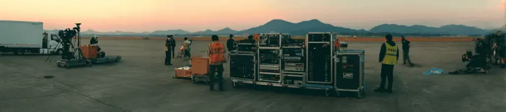 Several crew members in safety vests set up film equipment and large cases on an open tarmac at sunset, with mountains visible in the background—an inspiring scene of filming in Japan.