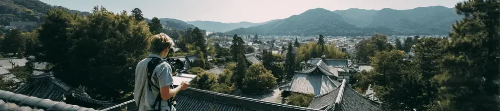 A person with a backpack stands on a rooftop, looking over traditional buildings surrounded by trees, capturing the beauty of Filming In Japan with a stunning town and mountains in the distance under a clear sky.