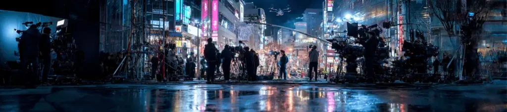 A nighttime city street in Japan bustling with film crew, cameras, and equipment, with colorful neon signs and lights illuminating the busy urban scene. A drone hovers above, capturing footage during the vibrant Filming In Japan experience.