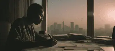 A person sits at a desk covered with papers, writing, while looking out a large window at a Johannesburg city skyline during sunset, capturing the inspiration behind filming in Johannesburg.