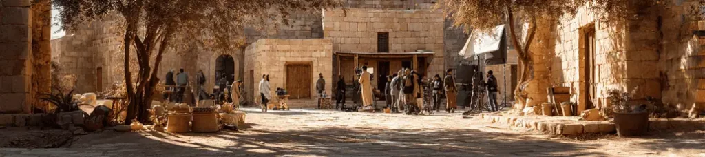 A film crew works on set, filming in Jordan’s dusty, sunlit courtyard surrounded by stone buildings, trees, and various props to capture the feel of an ancient or historical village.