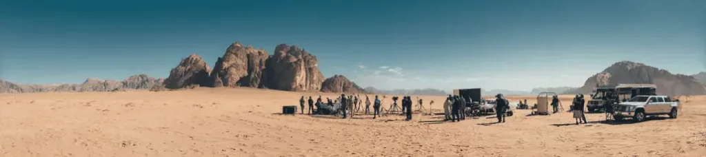 A film crew sets up equipment and vehicles in a vast desert landscape with rocky mountains in the background under a clear blue sky, capturing the magic of filming in Jordan.
