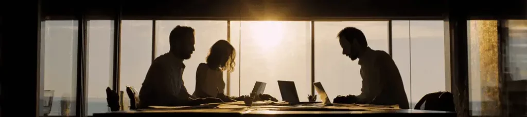 Silhouettes of three people working on laptops at a table in an office with large windows, as sunlight streams in from outside—a perfect setting for filming in Lebanon.