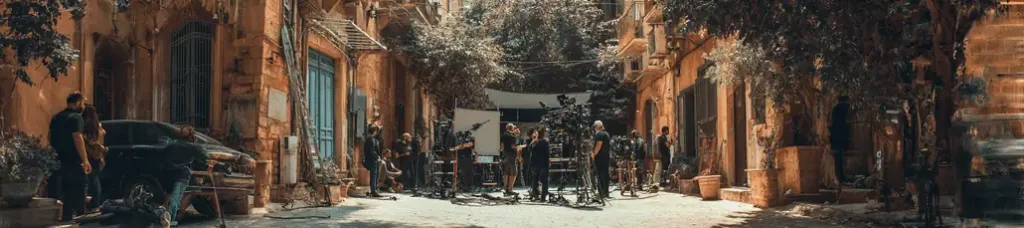 A film crew sets up cameras and equipment on a narrow, sunlit street lined with old buildings and trees, preparing for a shoot while people work and talk throughout the scene, capturing the vibrant energy of filming in Lebanon.