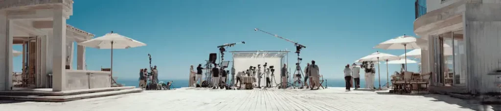 A film crew sets up cameras, lights, and equipment on a bright, sunny terrace overlooking the ocean in Lebanon, with people working under large white umbrellas and a clear blue sky above.