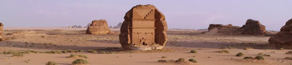 An ancient tomb carved into a large rock stands in a desert landscape of Saudi Arabia, with sandy ground, sparse vegetation, and distant rock formations under a clear sky—an evocative setting perfect for filming in Saudi Arabia.