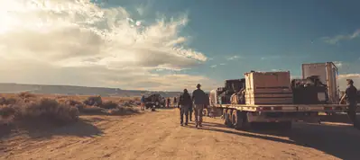 People walk along a dusty dirt road next to a trailer loaded with equipment under a bright sky with scattered clouds, capturing the spirit of filming in South Africa’s desert-like landscape.