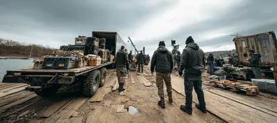 A group of people in winter clothing stand on a wooden dock, surrounded by equipment and supplies for filming in Norway. A truck loaded with items is nearby, with cloudy skies overhead and a body of water in the background.