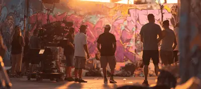 A film crew is filming in Miami outdoors at sunset, surrounded by vibrant graffiti-covered walls, with camera equipment visible and several people standing and talking in the warm, golden light.