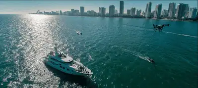 A large white yacht sails on blue water near several small boats, with a city skyline of tall modern buildings in the background under a bright, sunny sky—perfect for filming in Miami.
