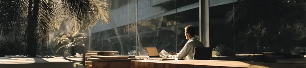 A person sits at a desk with a laptop and papers in a modern, sunlit office, facing large glass windows with views of palm trees and an urban building—an inspiring spot ideal for filming in Mexico.