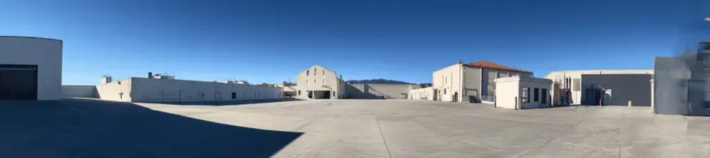 A wide, empty concrete lot surrounded by several white industrial buildings under a clear blue sky, with distant mountains visible in the background—an ideal location for filming in Mexico.