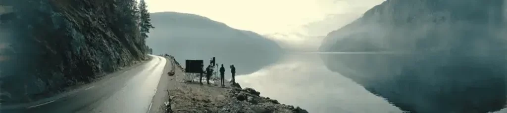 Four people stand by a lakeside road on a misty day, filming in Norway as calm water reflects mountains and trees; the scene is tranquil and atmospheric, with muted colors and low clouds over the lake.