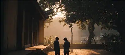 Two people stand and talk in the shade of large trees near a building at sunset, as warm sunlight filters through the branches—capturing the tranquil atmosphere of filming in Lebanon with long shadows cast on the ground.