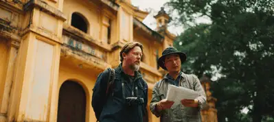 Two men stand in front of a yellow historic building; one wears a backpack and camera, and the other points at a paper, possibly reading a map or guide while filming in Vietnam.