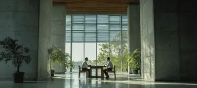 Two people sit across from each other at a small table in a spacious, modern room with tall concrete walls, large windows, and several potted plants, creating a calm, professional atmosphere perfect for filming in Vietnam.
