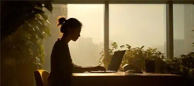 A person sits at a desk working on a laptop in a sunlit room, surrounded by plants and large windows with soft natural light streaming in—perfect for filming in Vietnam.