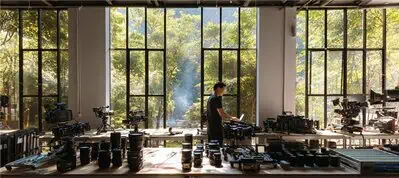 A person stands at a long table filled with camera equipment in a bright room with large windows, preparing for filming in Vietnam, while looking out onto lush green trees and sunlight streaming in from outside.