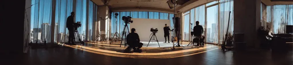 A film crew with cameras and equipment sets up in a spacious studio with large windows, sunlight streaming in, and a city skyline visible outside. Four crew members are preparing for a shoot, capturing the unique vibe of filming in Portugal.