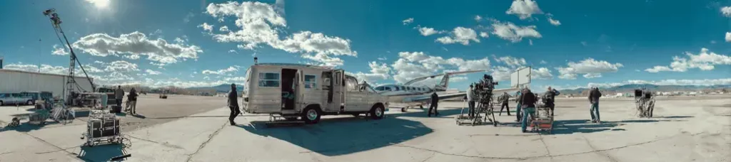A film crew sets up cameras, lights, and equipment around a small private airplane and a van on an airport tarmac under a blue sky with scattered clouds while filming in Portugal.