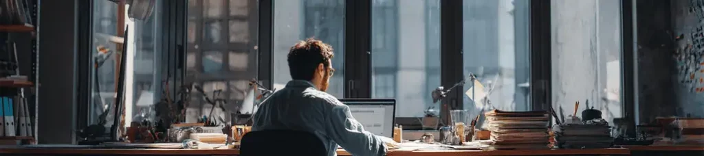 A man sits at a cluttered desk, working on a laptop in front of large windows. Natural light fills the room, which is lined with shelves and papers—an ideal setting for filming in Portugal amidst creative chaos.