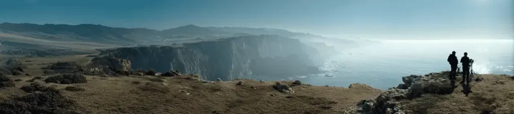Two people stand on a rocky cliff overlooking a vast, rugged coastline while filming in Portugal, with high cliffs, misty air, and the ocean below under a clear blue sky.