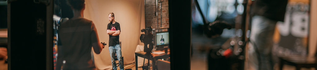 A man in a black t-shirt and jeans stands before a tan backdrop in a studio, facing another person. Studio lights, monitors, and a camera hint at an active Video Production Austria session in progress.