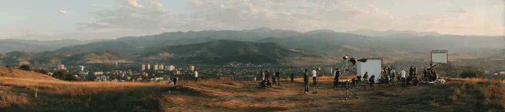 A wide view of a film set on a grassy hill overlooking a city, with mountains in the distance—filming in Romania—with scattered crew members, lighting equipment, and large screens visible.