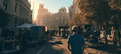 Film crew sets up equipment on a city street lined with historic buildings at sunset while filming in Madrid. People work among cameras, lights, and gear, with a silhouette in the foreground. The atmosphere is busy and cinematic.