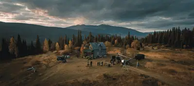 Aerial view of a film set in a rural area near Oslo, with a small house, crew members, equipment, and autumn trees under a cloudy sky with mountains in the background—perfect for filming in Oslo.