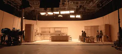 A film set designed as a kitchen is brightly lit with overhead lights. Several crew members stand near the kitchen island and counters, while cameras and equipment are visible around the set, capturing the excitement of filming in Boston.