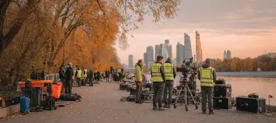 Film crew members wearing yellow vests set up cameras and equipment along a riverside path lined with autumn trees, filming in Moscow with a modern city skyline visible in the background.